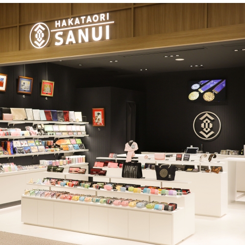 博多織の老舗「サヌイ織物」の店舗外観。木製のフレームで縁取られた入り口の上部にブランドロゴが光り、店内は黒い壁に白い陳列棚が映えるモダンな内装。中央と左右の棚には、色鮮やかなポーチや財布、バッグなどの博多織製品が多数展示されている。